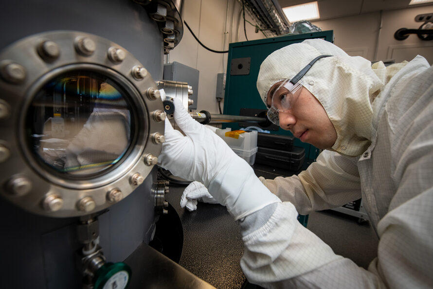 A scientist in a cleanroom suit, gloves, and safety glasses carefully examines a vacuum chamber, with their reflection visible in a round glass window.